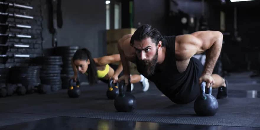 A man and a woman do push-ups with kettlebells in a gym, both focused and wearing athletic clothing, surrounded by fitness equipment and weights.