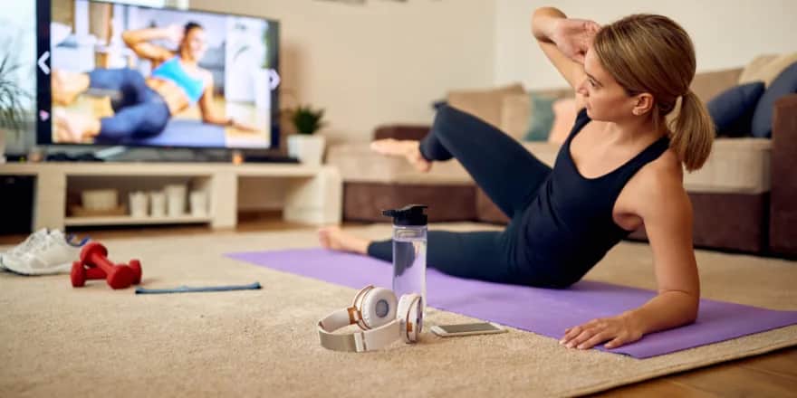 A woman exercises on a yoga mat in her living room, following a workout on TV. Dumbbells, a water bottle, headphones, and a smartphone are nearby. The room looks cozy with a sofa and shelves in the background.