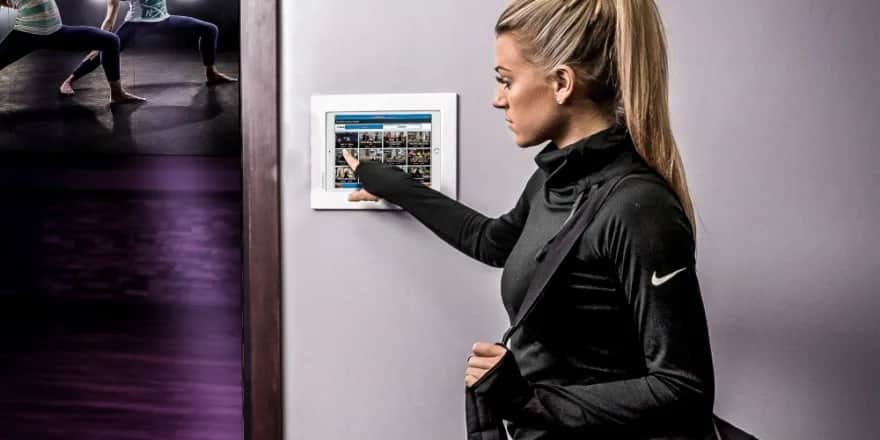 A woman with a ponytail and a gym bag uses a touchscreen panel on a wall, selecting from exercise or class options. In the background, people are doing yoga or stretching in a dimly lit studio.