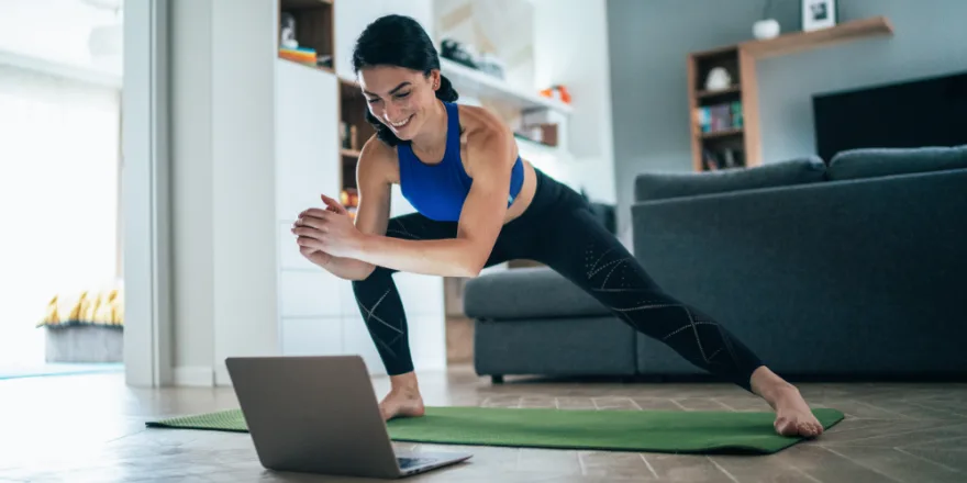 A woman in athletic wear stretches on a yoga mat in her living room while following an exercise video on a laptop. She smiles and leans to one side with hands clasped, with shelves and a couch in the background.