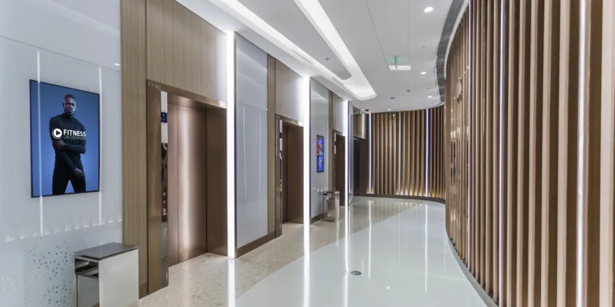 A modern hallway with elevators, wood paneling, and a polished floor. A digital sign on the wall displays a fitness ad with a man, and there is a trash can below it. The ceiling has built-in lighting.