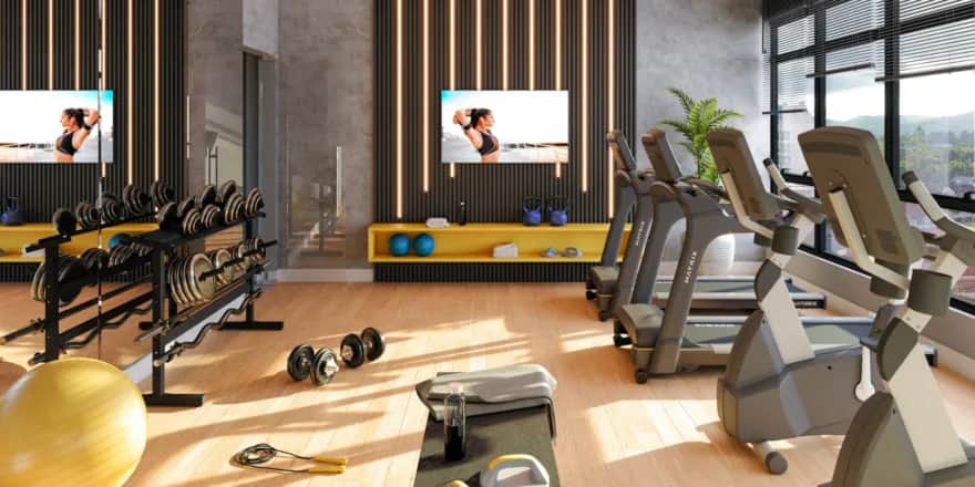 Modern gym with treadmills, stationary bikes, free weights, yoga mats, and a large yellow exercise ball. Sunlight streams through large windows, and two wall-mounted TVs display a woman exercising. The space is neat and bright.