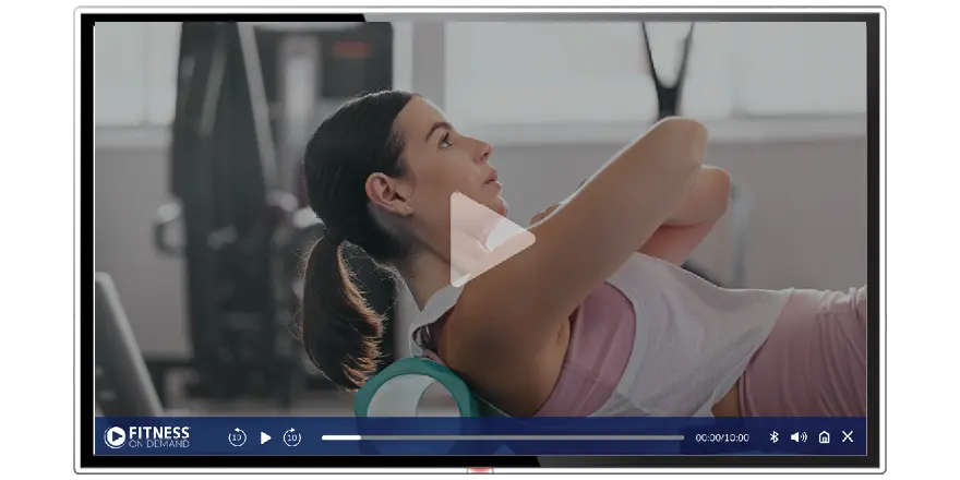 A woman in athletic wear rolls on a foam roller on a mat in a gym, shown on a video player screen labeled Fitness On Demand. Gym equipment is visible in the blurred background.