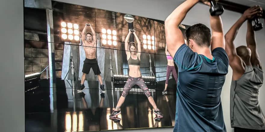 Two people exercise with kettlebells in a gym, following along with a workout video on a large screen showing three instructors demonstrating a kettlebell move.