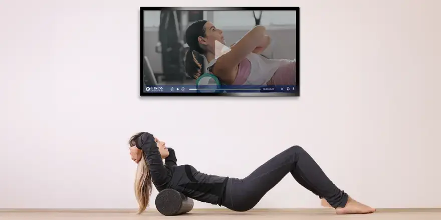 A woman in workout clothes lays on the floor using a foam roller under her upper back, while following a fitness video of a woman doing the same exercise on a wall-mounted touchscreen TV.