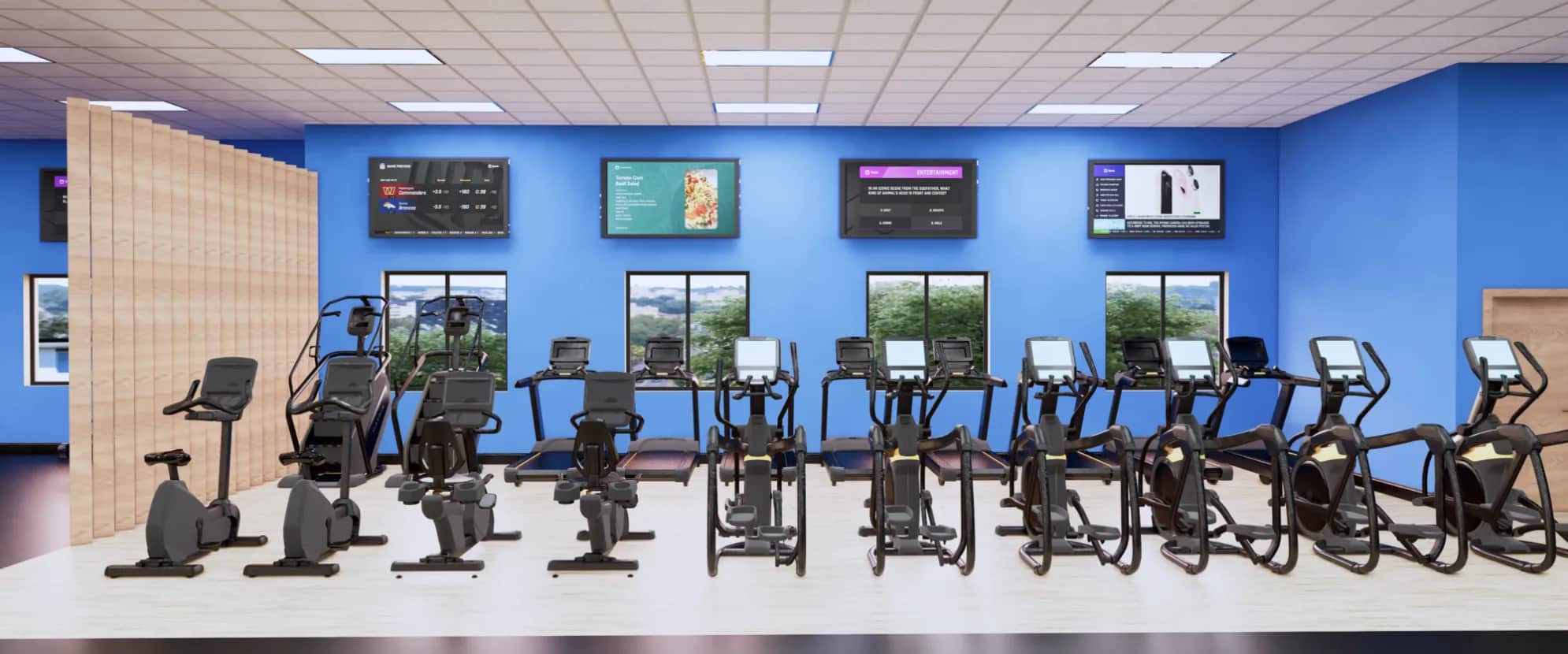 A row of stationary exercise bikes and elliptical machines is lined up in a brightly lit gym with blue walls, large windows, and TV screens mounted above the equipment.