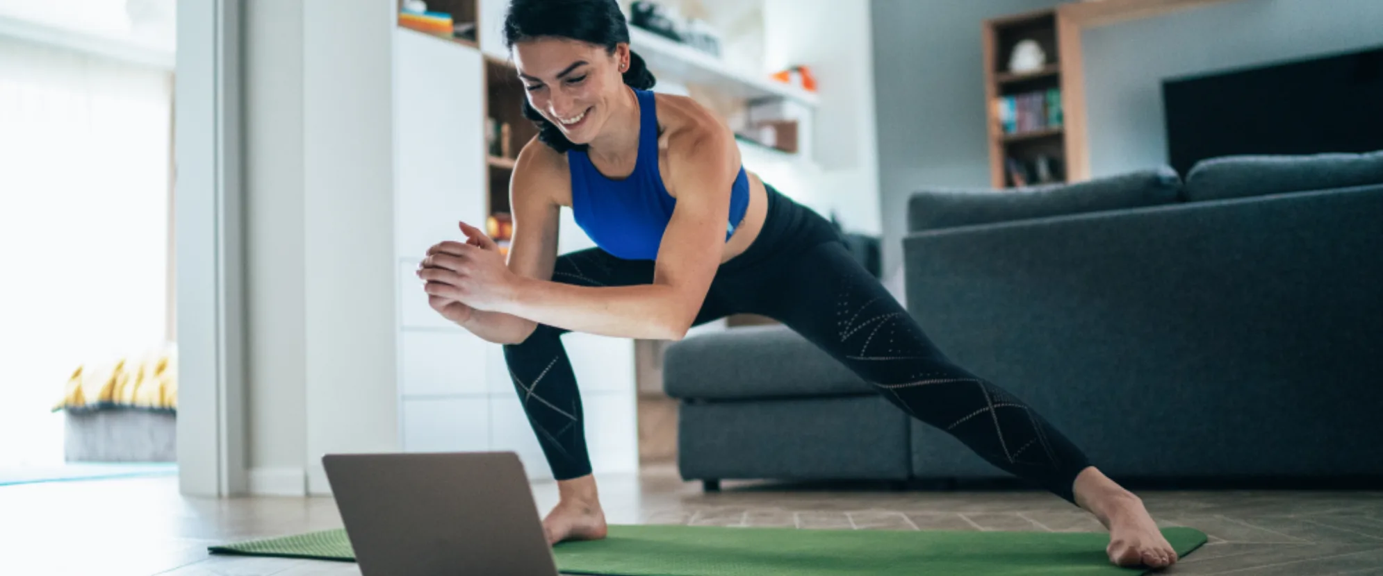 A woman in athletic wear stretches on a yoga mat in her living room while following an exercise video on a laptop. She smiles and leans to one side with hands clasped, with shelves and a couch in the background.