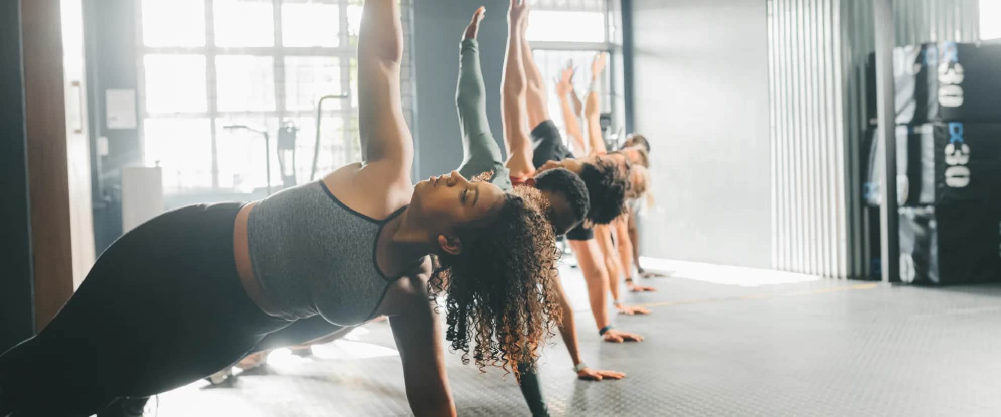 A group of people in athletic wear hold a side plank pose with one arm raised in a brightly lit gym, lined up on a rubber mat floor.