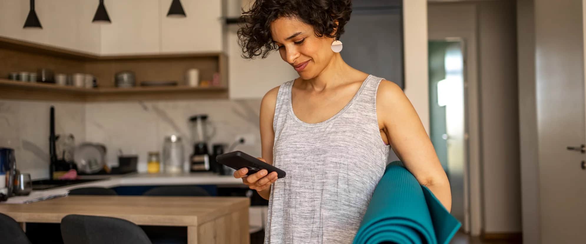 A woman with short curly hair holds a yoga mat under one arm and looks at her phone while standing in a modern kitchen. She is wearing a sleeveless dress and appears relaxed and focused.