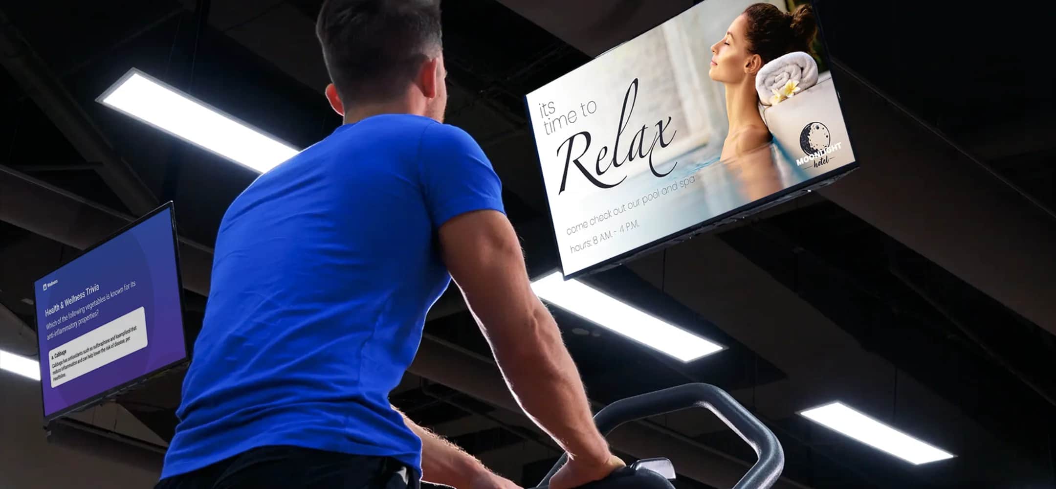 A man wearing a blue shirt uses a gym machine while looking up at a ceiling screen displaying an ad of a woman relaxing with towels and the text Its time to Relax. Fluorescent lights are visible overhead.
